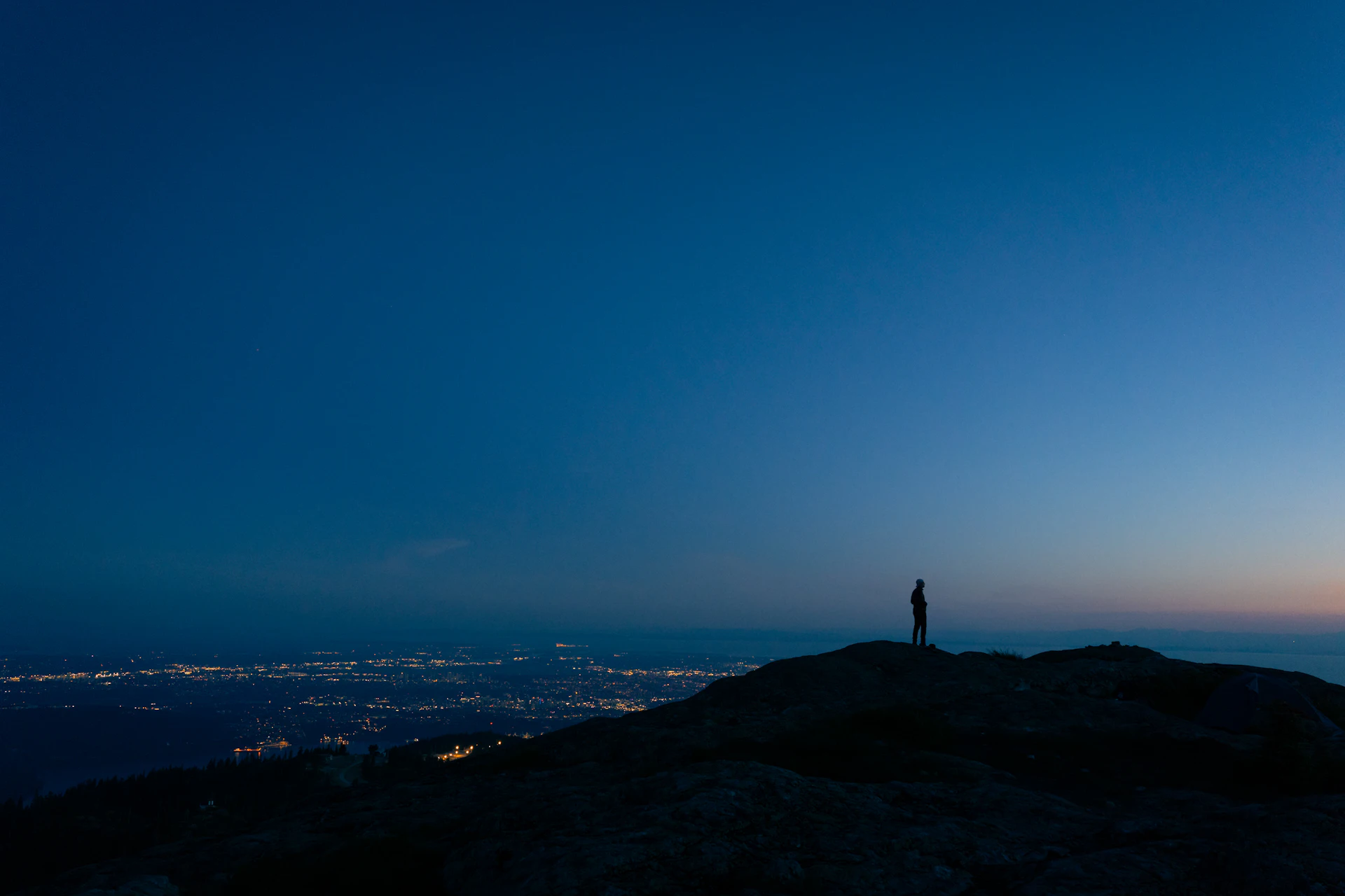 Lone person standing on a mountain ridge at dusk above distant city lights